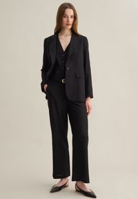 Veste noire ajustée avec revers crantés, assortie à un gilet boutonné et un pantalon droit. Des ballerines noires slim complètent la tenue.