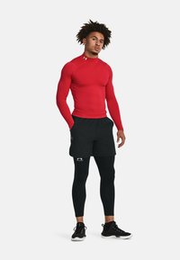 Rød langærmet sportsbluse med høj hals, kombineret med sorte shorts og sorte kompressionsleggings, der har Under Armour-logoer.