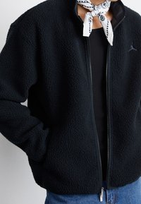 Czarna bluza polarowa z teksturowaną powierzchnią, zamkiem błyskawicznym i bocznymi kieszeniami. Na wysokości klatki piersiowej znajduje się małe logo. Noszona na czarnej bluzce.