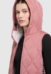 Pinker gepolsterter Weste mit Kapuze, die ein gestepptes Muster, einen Frontreißverschluss und schwarze Akzente an den Seiten aufweist, getragen über einem schwarzen Oberteil.