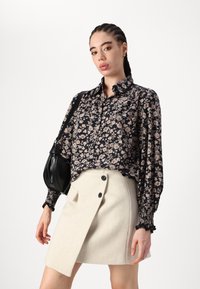 Blusa negra con estampado floral, de mangas largas y puños fruncidos, combinada con una falda envolvente beige que cuenta con un bolsillo lateral y botones negros.