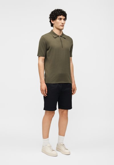 Άνδρας που στέκεται φορώντας ελαιί πράσινο πόλο με κοντά μανίκια και φερμουάρ, μαύρα κοντά παντελόνια με ρεβέρ, άσπρες κάλτσες και μπεζ αθλητικά παπούτσια σε λευκό φόντο.