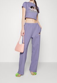 Gekürztes lila T-Shirt mit grafischem Design, kombiniert mit passenden lockeren Jogginghosen, einer rosa Handtasche und Sandalen mit grünen Fransenakzenten.