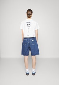 Camiseta blanca recortada con estampado en la parte trasera, combinada con pantalones cortos de mezclilla azul oscuro con dos bolsillos traseros. El modelo lleva zapatos azules y calcetines blancos.