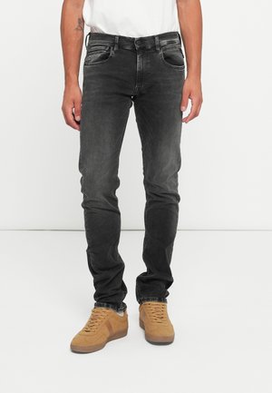 Man in vervaagde zwarte slim-fit jeans en tan suède sneakers, staand tegen een eenvoudige witte achtergrond.