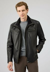 Zwarte leren jas met een gestructureerd ontwerp, twee ritszakken op de borst, zijzakken en een geribde kraag. Draag over een grijze shirt.