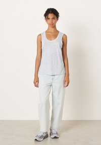 Mlada žena s kovrčavom kosom, odjevena u svijetlosivu majicu bez rukava, široke svijetloplave traperice i sive tenisice, stoji naslonjena na bijeli zid.