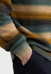 Meerleurige gebreide trui met horizontale strepen in teal, goud en bruin. Geribbelde manchetten en zoom, zachte textuur, gelaagde uitstraling.