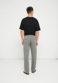 T-shirt nera a maniche corte con polsini bianchi, abbinata a pantaloni grigi chiari con tasche posteriori. Modello di spalle su uno sfondo neutro.