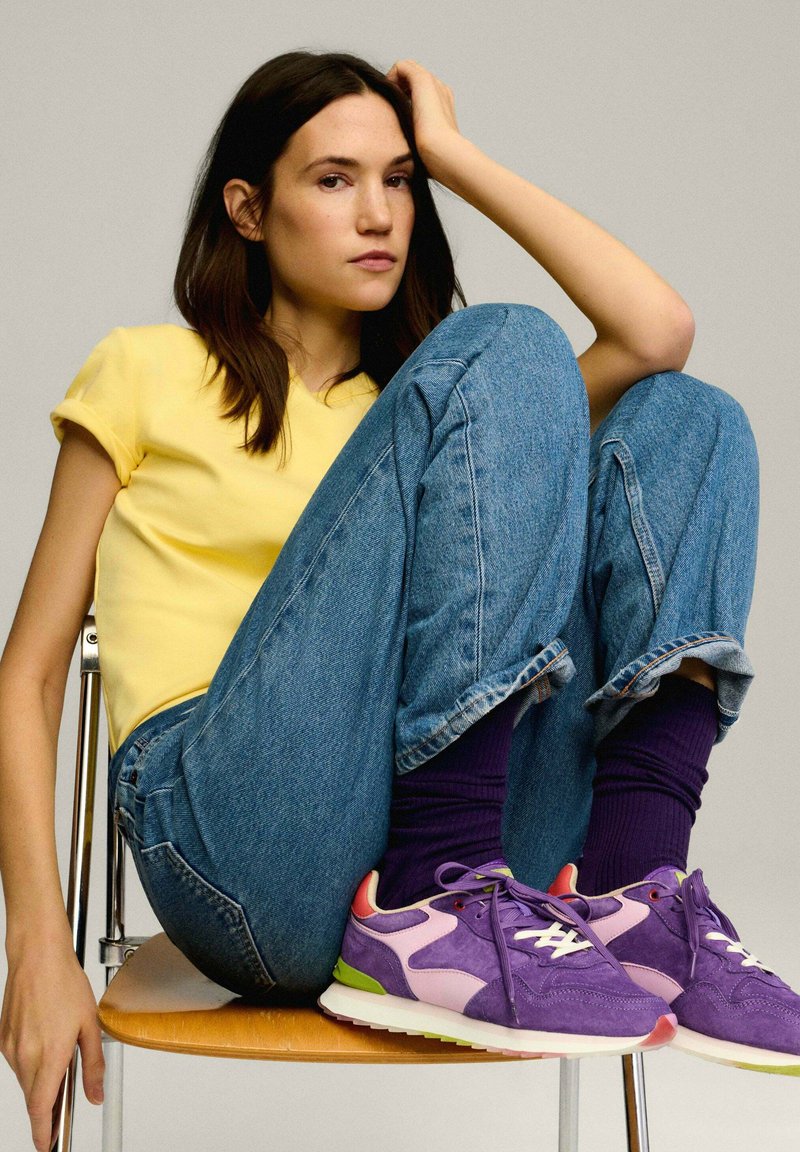 Baskets en suède violet avec des accents roses et verts ; portées avec un pantalon en denim bleu roulé et des chaussettes violettes, assis sur une chaise.