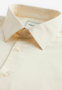 Chemise à manches courtes couleur crème avec un tissu texturé, un col pointu et deux boutons à l'avant. Étiquette de la marque visible à l'intérieur du col.