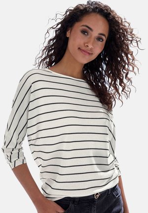 Gestreiftes Strickoberteil in Weiß und Schwarz, mit Rundhalsausschnitt, langen Ärmeln und einer lockeren Passform, kombiniert mit dunklen Jeans und einem Gürtel.