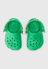 Um par de Crocs verdes com alças florais ajustáveis e furos de ventilação, vistos de cima sobre um fundo branco.