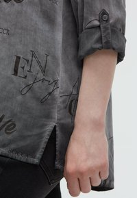 Rechter Arm mit hochgekrempeltem grauen Hemdsärmel, der schwarzen, geschwungenen Text und einen Seitenschlitz aufweist, kombiniert mit schwarzen Hosen.