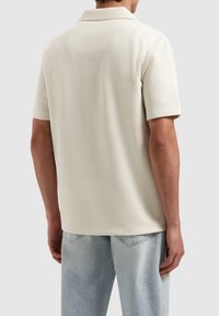 Korte mouwen, textuur crème poloshirt met een kraag; gecombineerd met lichtblauwe jeans. De stof heeft een subtiel, verheven patroon.