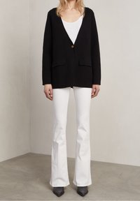 Black cardigan with a single bronze button, two front pockets, worn over a white tank top and paired with white flared pants and black heels.