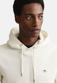 Sweat à capuche blanc avec cordon de serrage, arborant un petit logo brodé sur la poitrine. Tissu doux à la texture lisse et poignets côtelés.