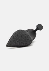 Svart gummiskor med spetsig tå, texturerad ovandel och märkes-sula med upphöjda bokstäver. Minimalistisk design med slät yta.