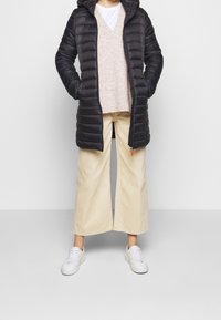 Schwarze isolierte Pufferjacke mit Kapuze, mit strukturiertem Obermaterial, Reißverschluss-Details, getragen über einem beigen Pullover und cremefarbenen, weit geschnittenen Hosen. Weiße Sneakers.