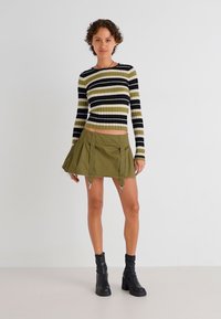 Pull court à rayures noires et olives en rib avec une jupe plissée verte et des accents métalliques, associé à des bottines noires.