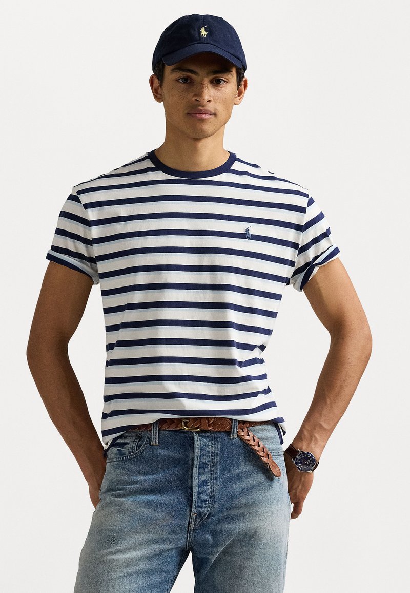 Randig bomulls-t-shirt med marinblå och vita ränder, ribbat runt halsen, broderad logotyp på bröstet, tillsammans med blekblå jeans och ett brunt bälte.