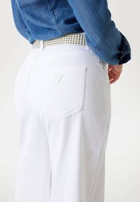 Osoba nosząca białe dżinsy z małym trójkątnym logo marki na tylnej kieszeni oraz zdobionym białym paskiem, w zestawieniu z niebieską koszulą dżinsową.