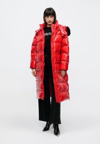 Rode gewatteerde lange jas met een glanzende afwerking, voorzien van een met bont gevoerde capuchon, ritssluiting aan de voorkant en een horizontaal stikselpatroon. Draagt over zwarte kleding.