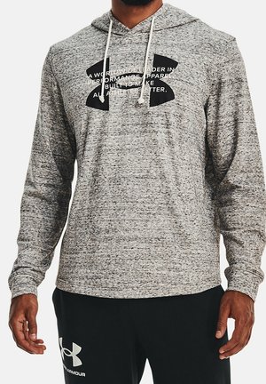 Mężczyzna ubrany w szarą bluzę Under Armour z kapturem na troczkach oraz czarne spodnie z logo Under Armour na lewej udzie.