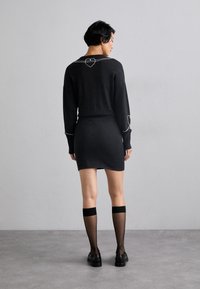 Pulover negru tricotat cu detalii cusute în formă de inimă pe spate, asortat cu o fustă neagră mulată și ciorapi netransparenti până la genunchi.