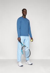 Sudadera azul, pantalones de chándal azul claro, zapatos blancos. Sosteniendo una raqueta de tenis y una pelota amarilla. Diseño simple, vestimenta atlética casual.