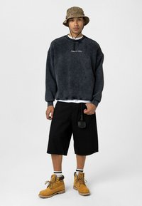 Donkergrijze oversized fleece sweatshirt met de tekst "De Toekomst Is Hier", zwarte shorts en gele veterschoenen voor werk. Accessoire bevestigd aan de shorts.