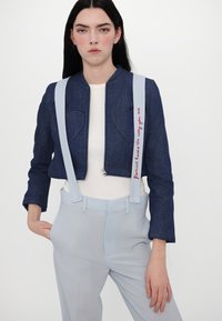 Jaqueta de ganga com costuras em forma de coração, suspensórios azuis e texto bordado em vermelho; combinada com calças azuis claras e uma blusa branca.