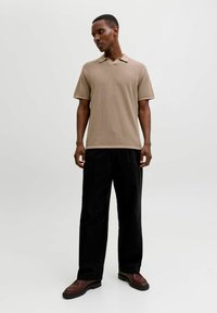 Homme debout, tête baissée, portant un polo en maille beige à manches courtes, un pantalon noir à jambes larges et des chaussures en cuir marron sur fond uni.