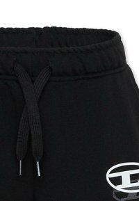 Pantalon de survêtement noir avec une taille élastique, un cordon de serrage ajustable, et un détail de logo blanc sur le côté gauche. Matière douce et texturée.