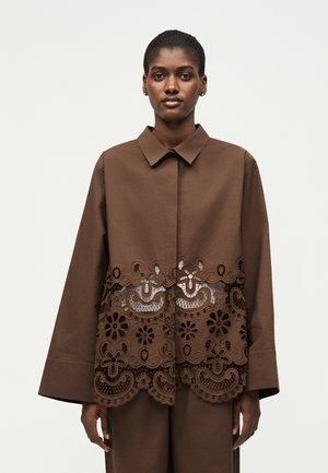 Nő barna hosszú ujjú felsőt visel, amelynek alsó felén virágos csipkemintázat található, és ehhez illő barna nadrággal van párosítva.