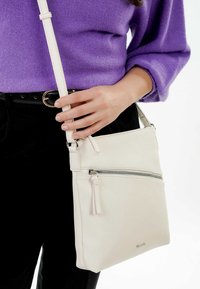 Borsa a tracolla bianca in pelle con tasca anteriore con zip, tracolla regolabile e superficie testurizzata. Indossata da una persona in un maglione viola.