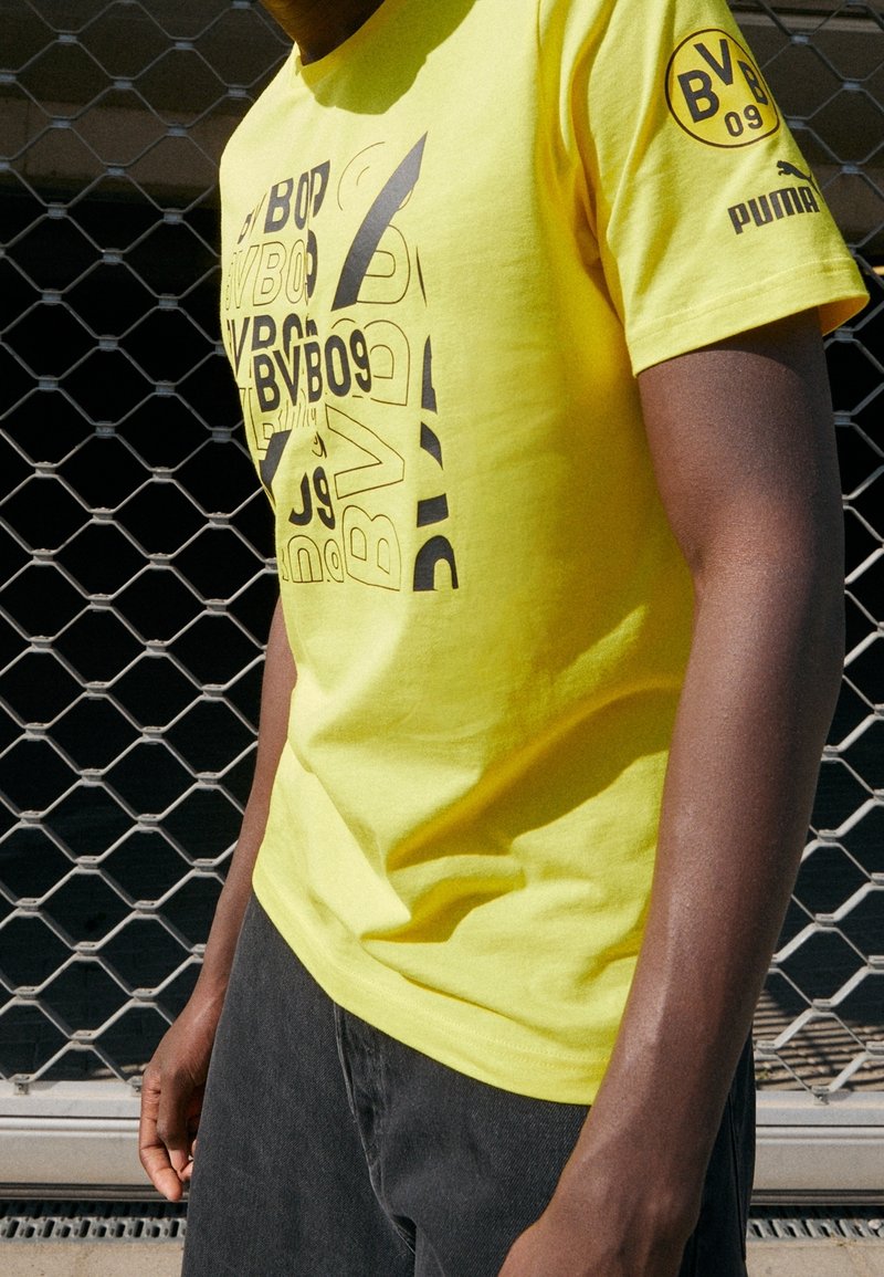 Camiseta de algodón amarilla con estampados de texto gráfico y logotipo en negro. Cuello redondo, mangas cortas, corte relajado. Lleva puesta con pantalones gris oscuro.