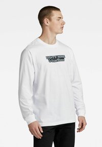 Hvit langermet t-skjorte med rund hals, prydet med fet grå tekst "EIGHTY NINE G-Star Raw" over brystet. Myk bomullsstoff.