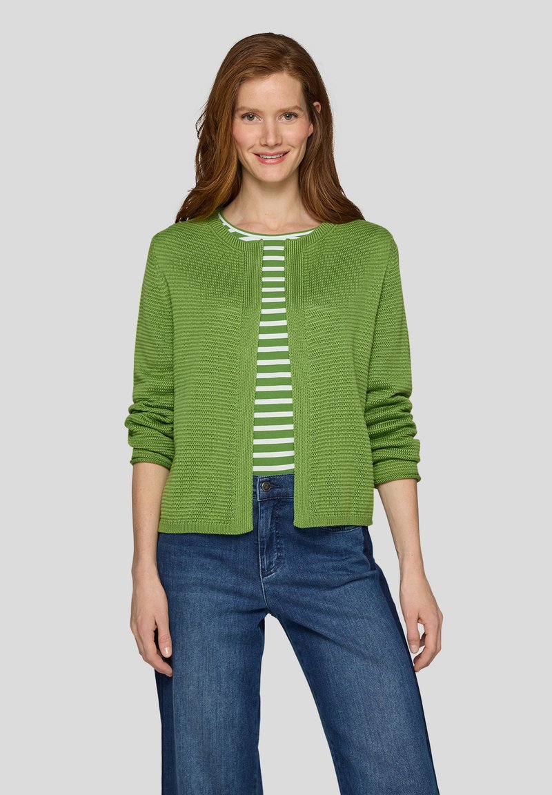 Frau mit langen roten Haaren, die einen grünen Strickcardigan über einem grün-weiß gestreiften Shirt und blauen Jeans trägt, steht vor einem schlichten Hintergrund.