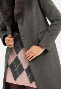 Grijze wollen jas met een donkere bontkraag, voorzien van twee knopen en zijzakken, gecombineerd met een roze argyle patroon rok.