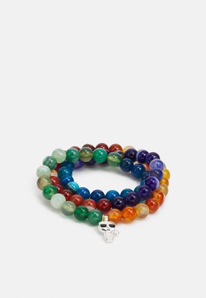 Bracelet multicolore en perles enroulé avec un charme tête de mort argentée portant des lunettes de soleil sur fond blanc.
