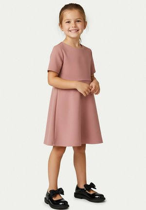 Lächelndes junges Mädchen steht, trägt ein kurzärmeliges staubrosa Kleid und schwarze Lacklederschuhe mit großen Schleifen.