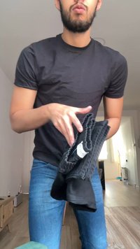 Junger Mann in schwarzem Hemd und blauen Jeans faltet schwarze Kleidung in einem hellen Raum mit Holzboden und Fensterlicht.