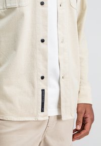 Beige overhemd met knopen, zwarte knopen, een patchlabel aan de onderkant en een contrasterend wit inner shirt dat zichtbaar is. Lichte stofstructuur.