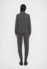 Tuta grigia composta da giacca con zip aderente e pantaloni a gamba affusolata. Strisce laterali viola in evidenza e polsini a coste. Tessuto liscio e strutturato.