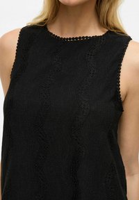 Must värvusega ilma varrukateta topp, millel on tekstuuriga kangas, esitleb lainelisi mustreid ja lainelist kaelust lisade jaoks.