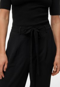 Vrouw draagt zwarte hoge taille broek met een stoffen riem en een aangesloten zwart topje met korte mouwen, handen ontspannen langs het lichaam.