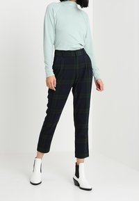 Lichtblauw langemouwtop met hoge hals, gecombineerd met marineblauwe geruite broek. Model draagt witte enkellaarsjes met zwarte accenten.