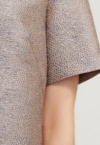 Krátký rukáv s texturovanou metalickou zlatou a levandulovou látkou na paži osoby na neutrálním pozadí.