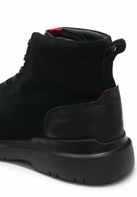 Botte montante en suède et cuir noir avec semelle épaisse texturée, languette au talon, accent rouge à l’intérieur du col et « HUGO » embossé sur le côté.